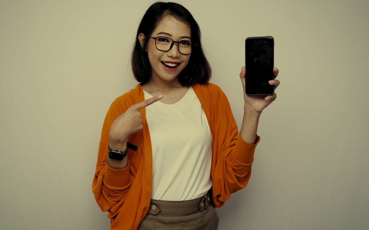 A smiling woman in a casual outfit holding up a smartphone, suggesting the integration of mobile technology in everyday life.